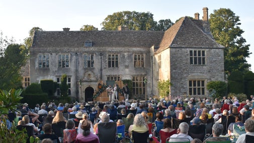 A large crowd watching a performance of A Midsummer Night's Dream in Avebury Manor Garden, with the 16th Century Avebury Manor house in the background behind the stage.